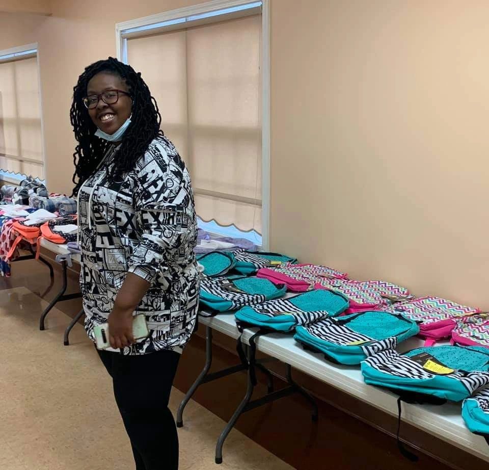 A WRWG volunteer stands beside a table filled with colorful donated backpacks